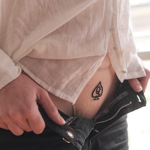 Könnte beinhalten: Schwarze Jeanshose mit offenem Reißverschluss und weißem Hemd. Ein Tattoo eines schwarzen Pik mit einem "Q" ist auf der Haut zu sehen.