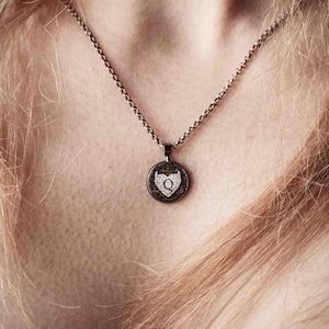 Può includere: Una collana color argento con un ciondolo rotondo. Il ciondolo presenta uno sfondo scuro con un disegno a forma di cuore bianco e la lettera "Q" bianca al centro. La collana è indossata intorno al collo.