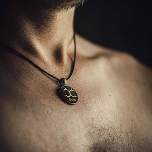Op de afbeelding: Een ketting met een ovale hanger en een zwart koord. De hanger heeft een goudkleurig ontwerp en de letters "KSW" aan de onderkant. De ketting wordt op de huid van een persoon gedragen.