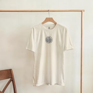 Puede incluir: Camiseta color crema con mangas cortas colgada de una percha de madera. La camiseta presenta un gráfico circular en la parte delantera con el texto "THE FOOTBALL PAPERS" en azul oscuro. La camiseta se muestra sobre un fondo neutro.