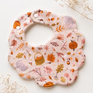 Könnte beinhalten: Ein Baby-Lätzchen mit gewelltem Rand in zartem Rosa, verziert mit einem wiederkehrenden Muster aus herbstlichen Motiven wie orangefarbenen Kürbissen, braunen Eicheln, bunten Herbstblättern, einem geflochtenen Korb und kleinen Igeln, mit Druckknöpfen.