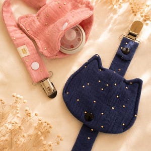May include: Two fabric pacifier holders shaped like cat heads, one in dusty pink and one in navy blue. Both feature gold metallic speckles, a metal clip, a white or black snap button, and a small fabric tag with the text TAKIO MAMA.