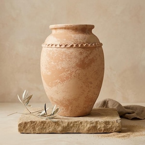 Könnte beinhalten: Eine Vase aus Terrakotta mit einem breiten, bauchigen Körper und einem schmalen Hals. Die Oberfläche weist eine gesprenkelte, verwitterte Textur in hellbraunen und beigen Tönen auf. Ein dekoratives, erhabenes Band in Seiloptik umgibt den oberen Teil.