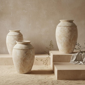 May include: Three beige, textured ceramic vases of varying sizes. Each vase has a rounded body and a beaded detail around the neck. The vases are displayed on a neutral-toned surface, creating a minimalist and earthy aesthetic.