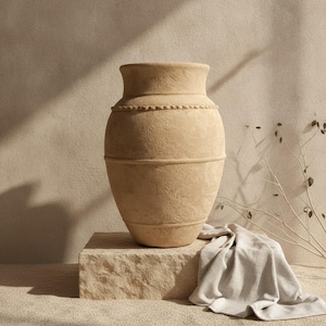 Peut inclure: Un grand vase en céramique beige à la texture marquée, doté d'un corps large et arrondi ainsi que d'un col étroit. Le vase présente une bande horizontale en relief sur la partie supérieure et une finition de surface subtilement irrégulière.