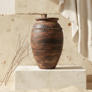Puede incluir: Un jarrón de madera redondeado de color marrón oscuro con una abertura ancha y una banda horizontal tallada y texturizada que rodea la sección superior. La superficie muestra patrones de veta de madera visibles con tonos marrones variados.