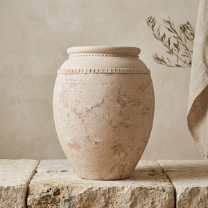 May include: A beige terracotta vase with a weathered, textured finish and a wide, rounded body. The neck features a decorative band with small, raised bead and vertical line details. The surface has a mottled, uneven appearance in light tan.