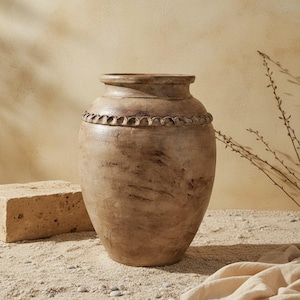 Peut inclure: Un grand vase en céramique beige avec une surface texturée. Le vase a un corps large et arrondi et un col plus étroit avec une bande décorative et nervurée. Le fond comprend une surface sablonneuse et un fond beige.