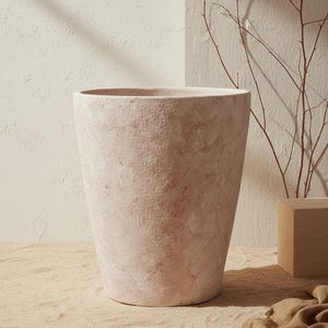 Könnte beinhalten: Ein großer, hellrosa Pflanztopf mit strukturierter Oberfläche. Der Pflanztopf hat eine breite Öffnung und eine konische Form. Der Hintergrund umfasst eine neutralfarbene Wand und einen kleinen Holzblock mit Zweigen.