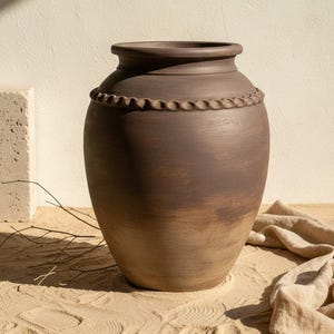 Op de afbeelding: Een grote, bruine keramische vaas met een brede body en een smalle hals. De vaas heeft een decoratief touwachtig detail rond de bovenkant. De vaas staat op een zanderige ondergrond, met een neutrale achtergrond.