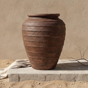May include: A large, dark brown, rustic earthenware vessel with a wide, rounded body and a thick, prominent rim. The exterior surface features a textured, weathered finish with horizontal ridges encircling the entire circumference of the pot.