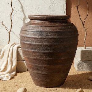 May include: A large, brown, textured ceramic vase with a wide base and a narrow neck. The vase has horizontal ridges and a dark, earthy tone. The background includes sand, stones, and decorative branches, suggesting a natural, rustic aesthetic.