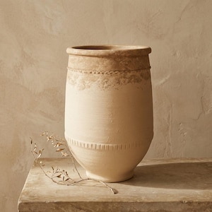 Può includere: Un vaso alto in terracotta beige con un bordo largo e arrotondato e una base affusolata. La parte superiore presenta una finitura ruvida e usurata con una fascia decorativa, mentre il corpo inferiore mostra un sottile motivo verticale inciso.