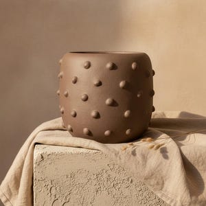 Puede incluir: Una maceta de cerámica marrón mate con forma cilíndrica redondeada que presenta un patrón repetitivo de pequeños bultos circulares en relieve en toda su superficie exterior. La maceta tiene una abertura superior ancha y un acabado uniforme.