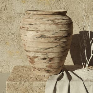Pode incluir: Um vaso grande de cerâmica bege texturizada com faixas horizontais. O vaso está sobre um bloco de pedra, com um pano de cor neutra drapeado sobre ele. Um galho branco está à direita do vaso.