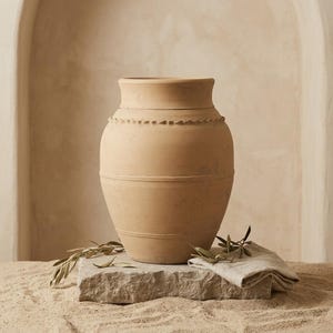 May include: A light tan terracotta vase with a wide, rounded body and a flared rim. The exterior features horizontal incised lines and a decorative scalloped band around the upper section. The surface has a matte, earthy texture throughout.