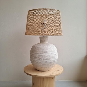 Puede incluir: Lámpara de mesa decorativa con pantalla tejida y base esférica de cerámica blanca texturizada. La lámpara está sobre una pequeña mesa auxiliar redonda de madera. El diámetro de la base es de unos 30 cm.