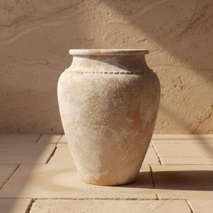 Puede incluir: Un jarrón grande de terracota beige con una base ancha y un cuello estrecho. El jarrón tiene una superficie texturizada y una línea punteada decorativa alrededor del cuello. El jarrón se exhibe sobre una superficie de piedra clara.