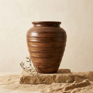 Pode incluir: Um vaso de madeira castanho-escuro com um corpo largo e arredondado, apresentando uma série de ranhuras horizontais em todo o exterior. O vaso tem um rebordo superior espesso e plano, com um acabamento polido que realça o veio da madeira.