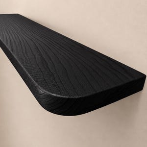 May include: A wall-mounted floating shelf featuring a solid black finish with a prominent wood grain texture. The shelf has a rectangular shape with one rounded corner, showcasing a smooth surface and clean edges for a simple display solution.