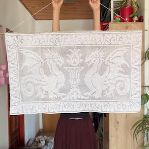 Könnte beinhalten: Ein rechteckiger weißer Häkel-Wandbehang mit einem detaillierten Filetspitzenmuster aus zwei symmetrischen Drachen, die einem zentralen Blumenmotiv gegenüberstehen. Das Stück hängt an einem dünnen Holzstab an einer weißen Schnur.
