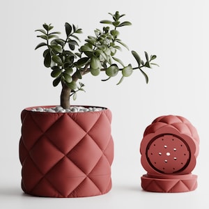 May include: A red quilted planter with a jade plant and white pebbles. Beside it, a matching red decorative piece with a floral design. The planter has a diamond pattern.