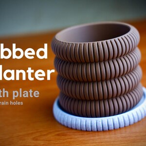 May include: Four brown, ribbed, circular planters stacked on a white ribbed plate. The planters have a ridged design. The text "ribbed planter with plate and drain holes" is visible. The planters are designed for plants.