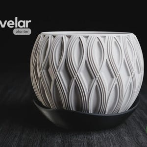 May include: A white spherical planter with a textured, repeating geometric pattern of vertical, curved lines and diamond shapes. The planter sits inside a black, curved saucer. The text "velar planter" appears in the upper left corner.