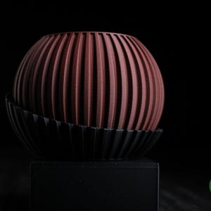 Può includere: Oggetto decorativo sferico con scanalature verticali in una tonalità marrone rossastro, appoggiato su una base nera curva. L'oggetto è posto su uno sfondo scuro, evidenziando la sua consistenza e forma. La base poggia su un quadrato nero.