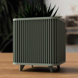 May include: A square, sage green planter with vertical slat detailing. The planter has short, tapered legs and contains a dark green succulent plant. The design is modern and minimalist, suitable for indoor or outdoor use. The planter sits on a wooden surface.