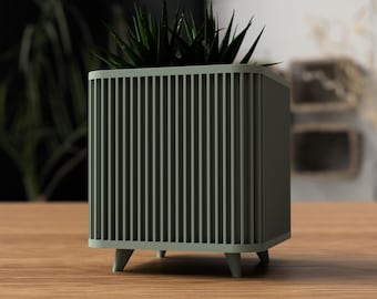 May include: A square, sage green planter with vertical slat detailing. The planter has short, tapered legs and is filled with dark green, spiky plants. The planter sits on a wooden surface, with a blurred background.