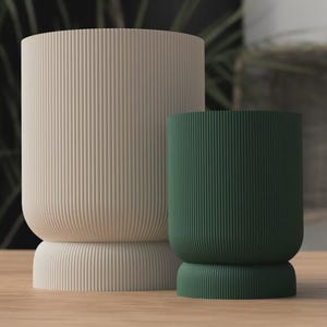 Puede incluir: Dos maceteros cilíndricos con textura acanalada vertical. Uno es de color beige claro y el otro verde oscuro. Ambos tienen una base y una parte superior redondeadas. Los maceteros están sobre una superficie de madera.