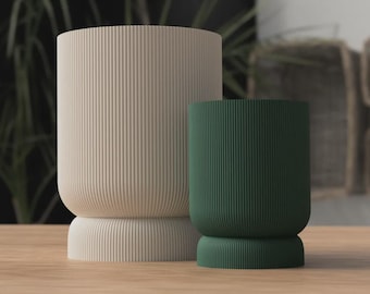 May include: Two cylindrical planters with vertical ribbed texture. One is a light beige color, and the other is a deep green. Both have a rounded base and top. The planters are on a wooden surface.