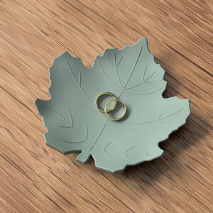 Puede incluir: Un plato decorativo en forma de hoja de color verde salvia con detalles de venas en relieve, que contiene dos anillos de metal dorado fino en el centro. El plato tiene un acabado mate suave y un borde lobulado, ofreciendo una superficie poco profunda.