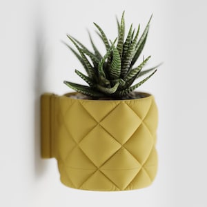 Puede incluir: Un macetero de pared amarillo con diseño de rombos y una planta suculenta. El macetero es de color amarillo brillante y tiene una textura acolchada. La suculenta tiene hojas a rayas verdes y blancas.
