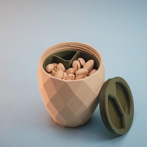 Puede incluir: Un cuenco para aperitivos de color beige claro con facetas y un patrón geométrico de diamantes. El cuenco tiene un divisor interno de color verde oliva y está lleno de pistachos. Una tapa circular verde oliva a juego descansa al lado.