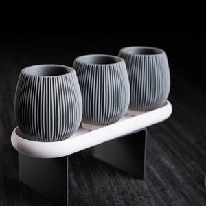 May include: Three grey, ribbed, rounded planters are arranged in a row on a white, oval-shaped tray supported by two dark grey, rectangular metal legs. The text h3liø design is visible in a small green circle in the bottom right corner.