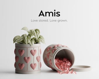 May include: A cylindrical planter with a light gray exterior, adorned with pink heart-shaped appliqués, holds a green leafy plant. Beside it, a matching overturned container spills pink decorative stones. The word "Amis" is displayed above the text "Love stored. Love grown."