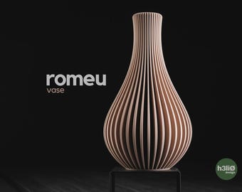 May include: A light tan vase with a vertical ribbed texture and a narrow neck. The text 'romeu vase' is printed in white and light tan on the left side. A small green circular logo with the text 'h3liø design' is visible in the bottom right corner.