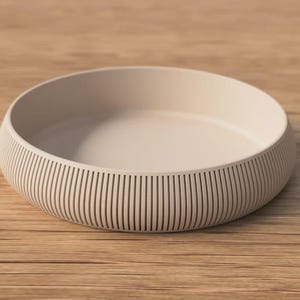 May include: A beige, circular bowl with a ribbed exterior. The bowl is placed on a wooden surface. The design of the bowl features vertical lines around the sides, creating a textured look. The bowl is empty.