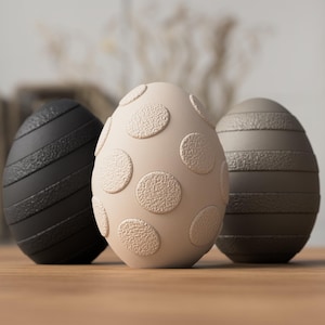 May include: Three decorative Easter eggs. The left egg is black with diagonal stripes. The central egg is beige with circular patterns. The right egg is grey with horizontal stripes. Each egg has a textured surface, suitable for Easter decorations.
