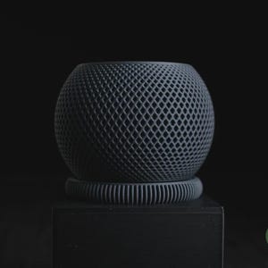 May include: A dark grey spherical planter with a diamond-shaped lattice pattern on its surface. The planter sits on a circular, ribbed base. A small green circular logo with the text h3liø design is visible in the bottom right corner.