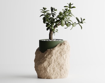 May include: A small green succulent plant with thick, rounded leaves potted in a dark olive green cylindrical container that is partially encased in a textured, light beige, rock-like base with a layer of small white pebbles on the soil surface.
