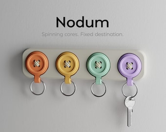 May include: A wall-mounted key holder featuring four colorful button-shaped hooks in orange, yellow, mint green, and lavender. Each button has a silver metal ring attached. The text "Nodum" and "Spinning cores. Fixed destination." is visible.