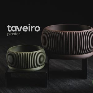 Peut inclure: Deux jardinières rondes à texture nervurée verticale, une vert olive et une marron foncé, reposant chacune sur un support en métal noir. Le texte "taveiro planter" apparaît en blanc, et un petit logo circulaire vert "h3liø design" est visible.