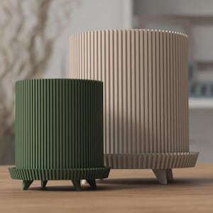 Puede incluir: Dos maceteros cilíndricos, uno verde oliva y otro beige, con detalles de ranuras verticales. Los maceteros tienen una base a juego con tres patas pequeñas. Los maceteros están hechos de un material sólido y son aptos para uso interior o exterior.