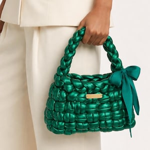 Può includere: Una borsa verde smeraldo intrecciata con un manico intrecciato e un fiocco abbinato. La borsa ha una forma rettangolare con una piccola placca color oro. La borsa è tenuta da una persona che indossa un abito bianco.