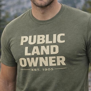 May include: Olive green t-shirt with the text "PUBLIC LAND OWNER" in a bold, off-white font. Below the text is "EST. 1905" in a smaller font. The shirt is short-sleeved and made of a soft material.