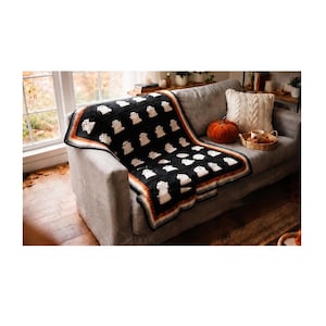 May include: A black crochet blanket featuring a repeating pattern of white ghost motifs. The blanket has a decorative border with stripes in shades of orange, brown, and cream, and is draped over a gray sofa with a textured white throw pillow.