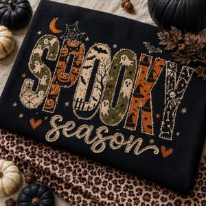 May include: A black sweatshirt featuring the text SPOOKY SEASON in a decorative applique style. The letters are filled with patterns of ghosts, pumpkins, bats, and skeletons. The word season is written in a cursive font with two small orange hearts.